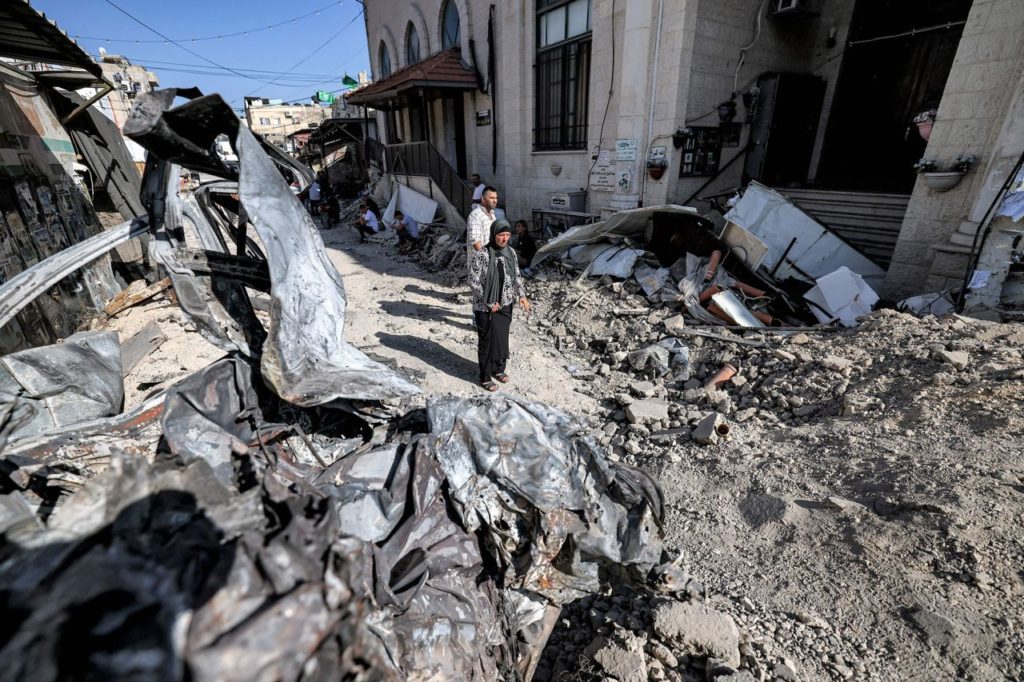 Palestinians are surrounded by rubble and the remains of a destroyed vehicle outside a mosque in Jenin, West Bank, on Wednesday, July 5, 2023 after a 2-day attack by Israel.