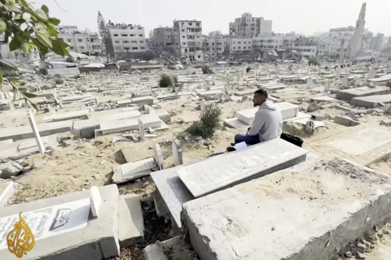 A Palestinian sits in a cemetery, looking at the rubble of buildings bombed by the Israeli military in Gaza