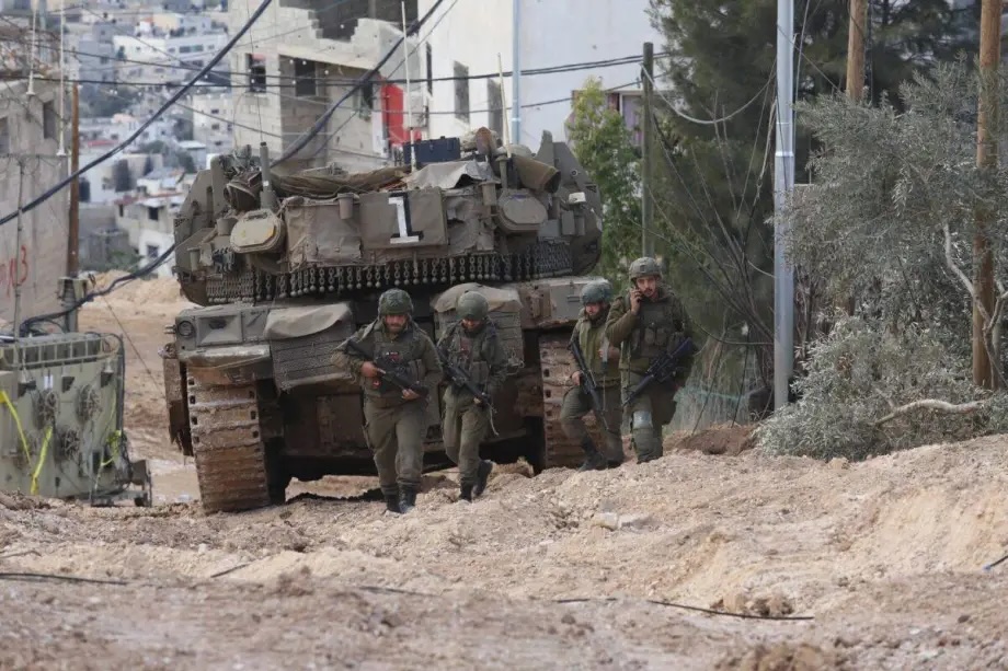 Israeli tanks and soldiers move through a muddy street during a raid at Jenin Refugee Camp in Jenin, West Bank, on February 24, 2025