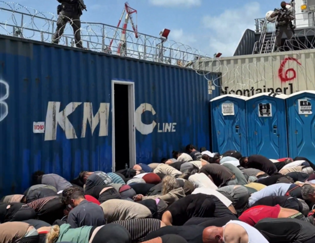 Bound flotilla activists kneeling on the floor, crowded, with soldiers standing above them on converted shipping containers, with machine guns drawn.